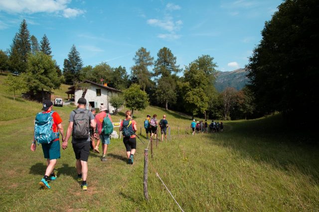 Group of travelers with backpacks walking through a grassy field toward a mountain house