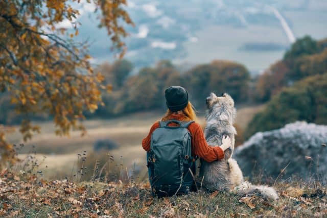 Woman with backpack and large dog sitting on a hill overlooking an autumn landscape