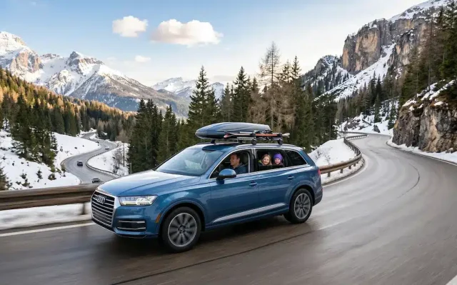 A family in a blue SUV with a ski roof box driving on a winding, snowy mountain road in the Italian Dolomites, representing a successful car rental experience for a trip to Paganella.