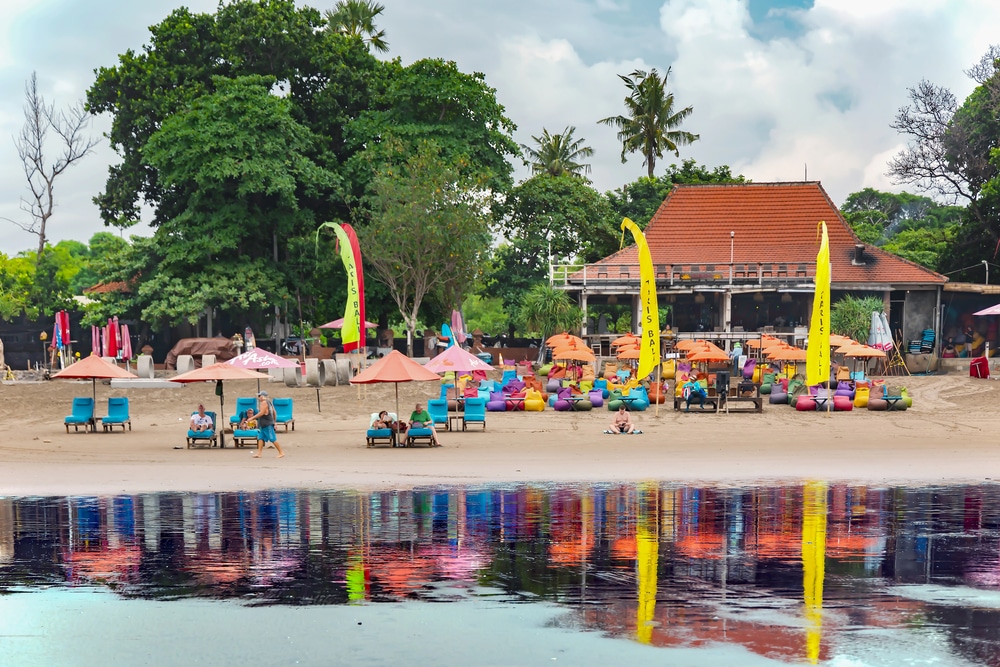 Colorful beach chairs, umbrellas, and beanbags set up on Seminyak Beach in Bali, Indonesia, with a traditional-style beach club in the background reflected in the shoreline water.