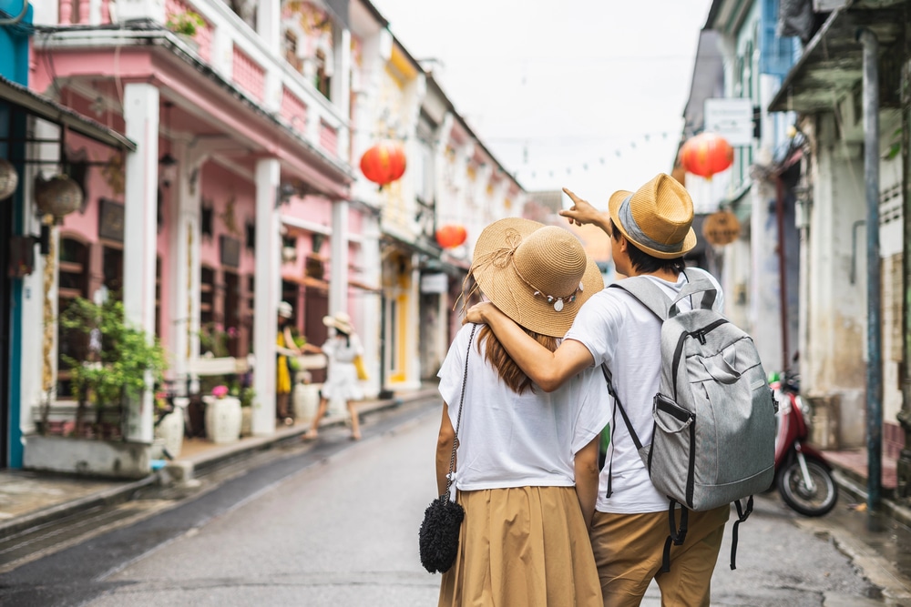 Rear view of a young couple with backpacks and straw hats walking down a colorful street with historic buildings and red lanterns, with one person pointing ahead.