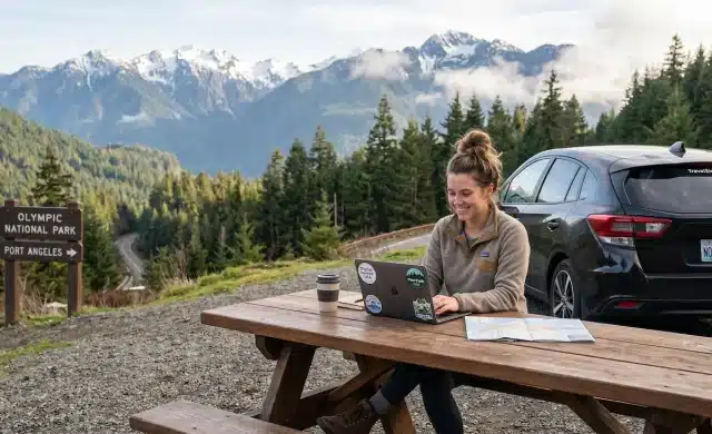 digital nomads working on laptops and reviewing a map at a picnic table in Olympic National Park, Washington, with a car parked nearby and snow-capped mountains in the background.