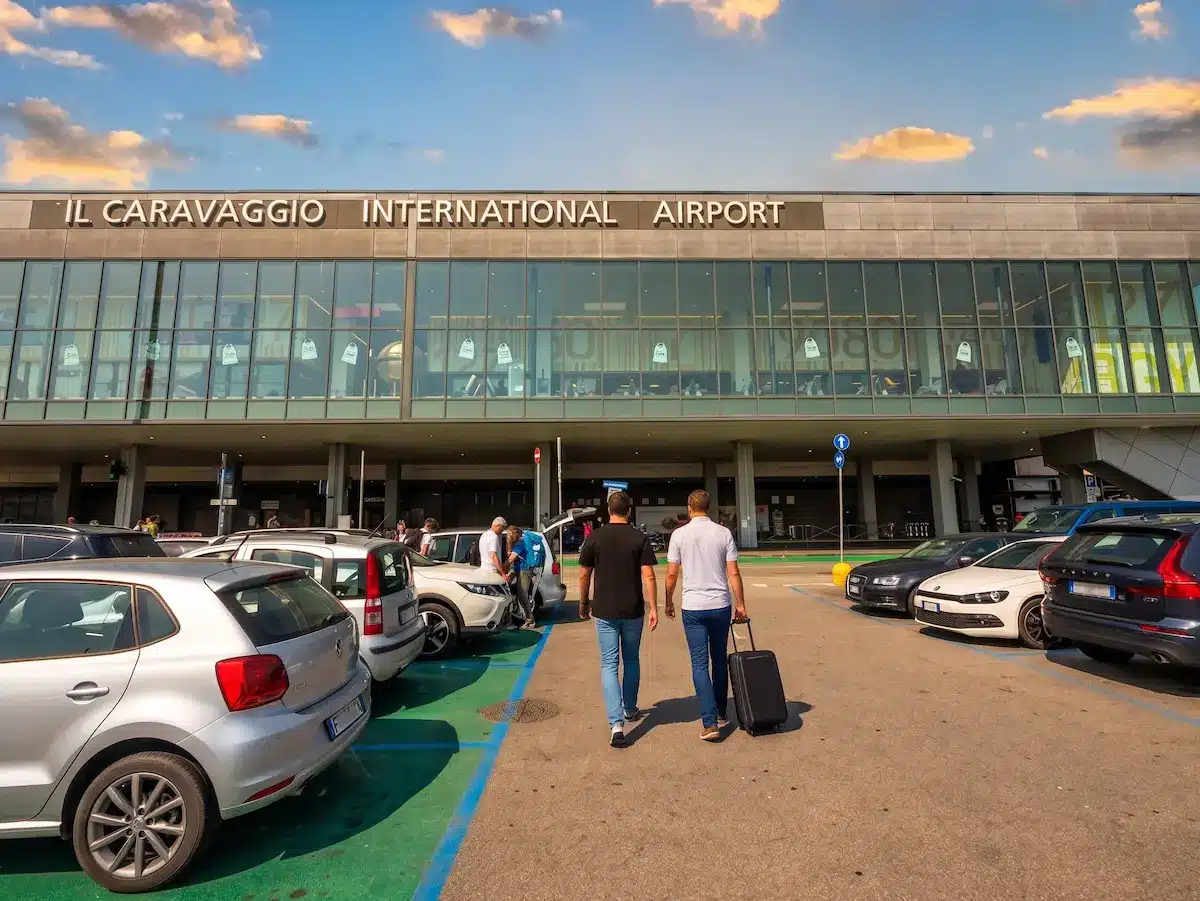 Two male travelers with suitcases walking across a sunny car rental parking lot toward the glass terminal building of Il Caravaggio International Airport in Bergamo, Italy.